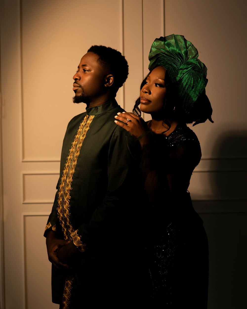 Two people pose dramatically in formal attire against a light wall, with one wearing a green headwrap and sparkly dress.
