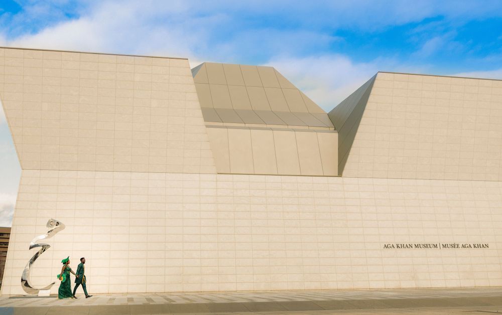 Two people walk past a modern beige building with angular walls under a blue sky.
