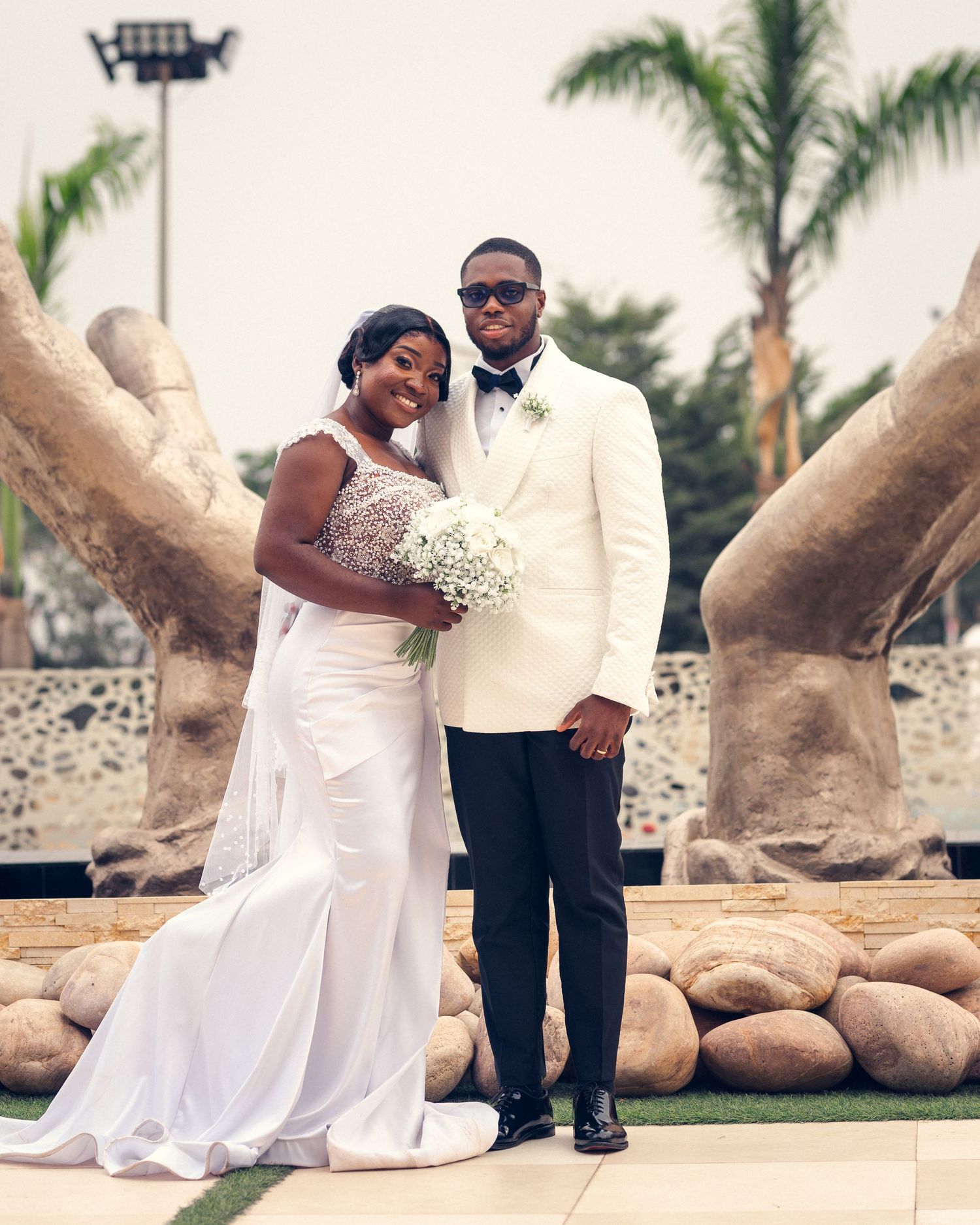 Bride in stunning lace gown holds bouquet next to groom in white tuxedo at outdoor venue.