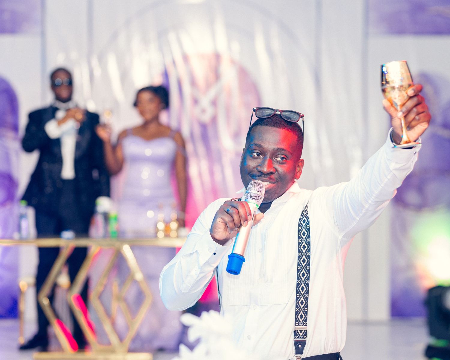 Wedding guest in suspenders and white shirt gives an energetic toast with a drink in hand.