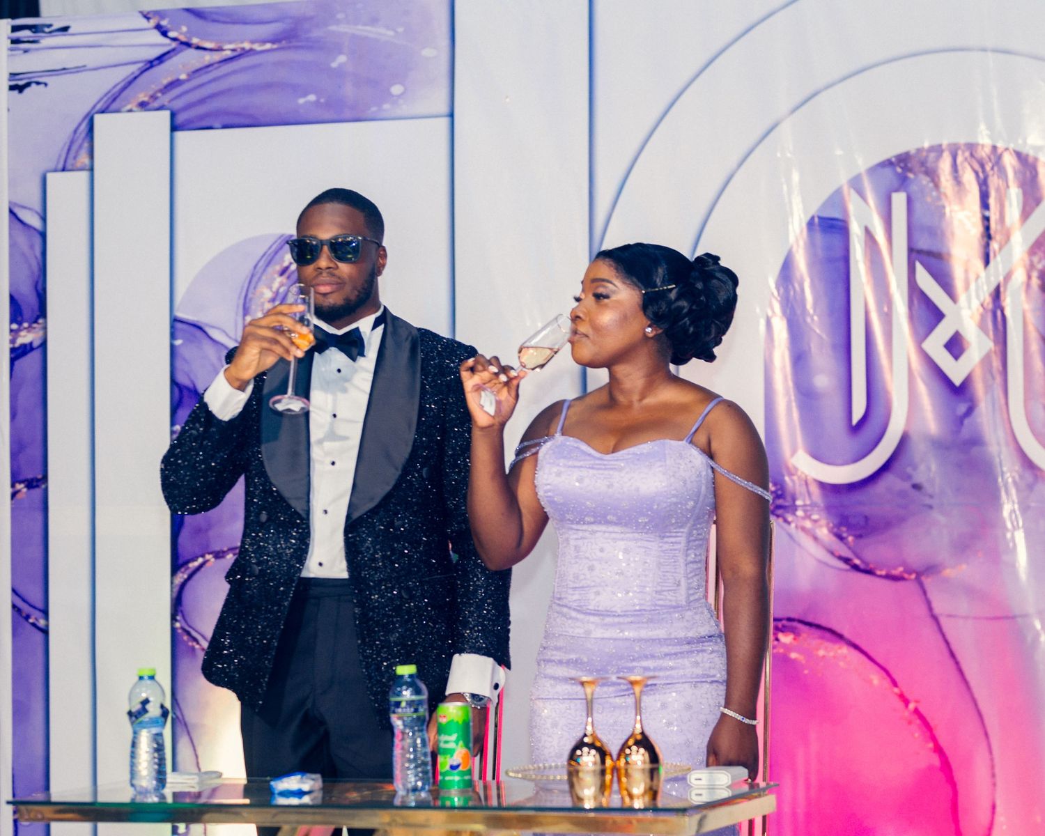 Couple in formal attire enjoying drinks at an elegant reception with neon lighting.