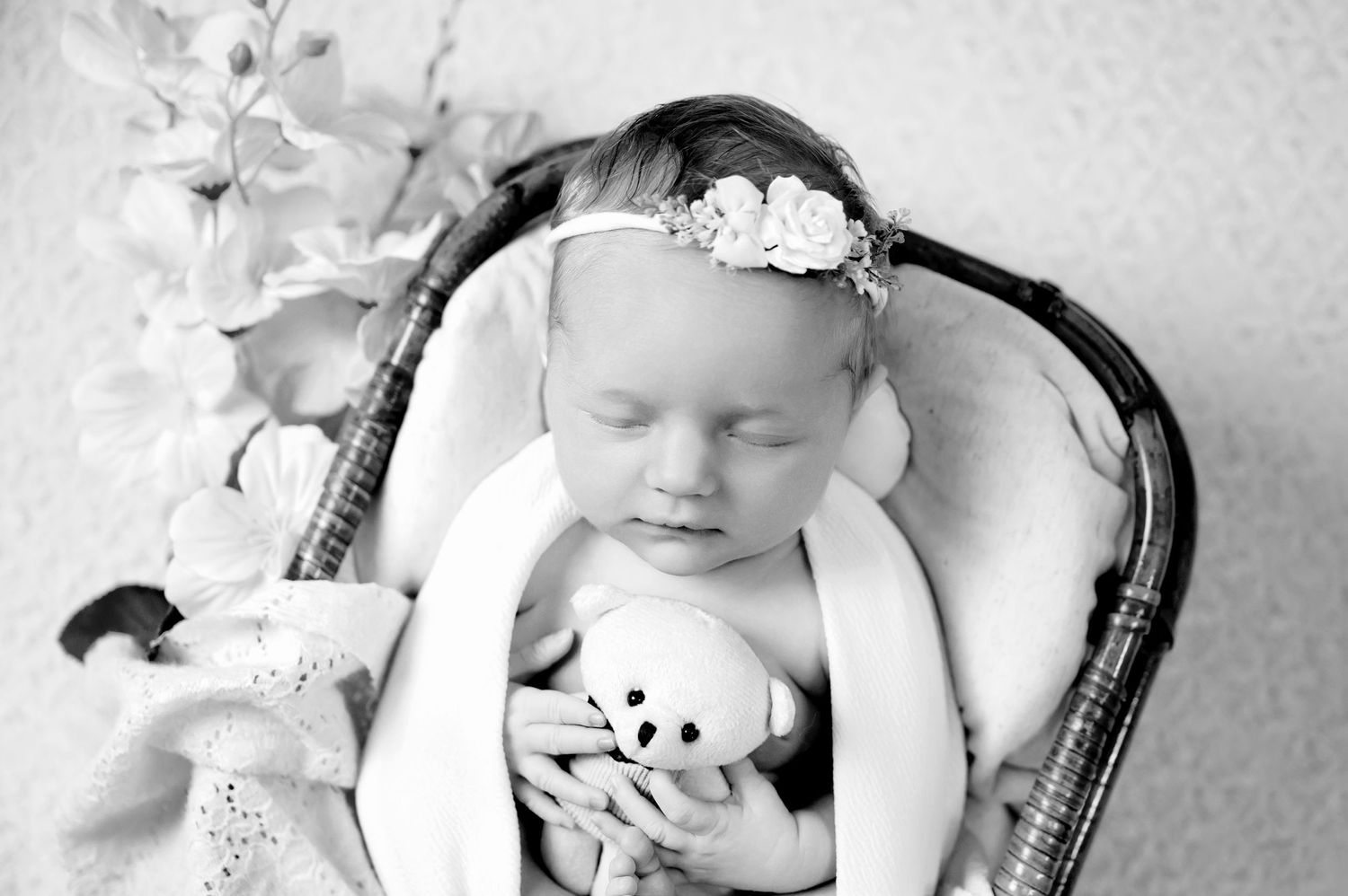 Black and white portrait of a baby lying in a wicker chair with a floral headband and peaceful expression.