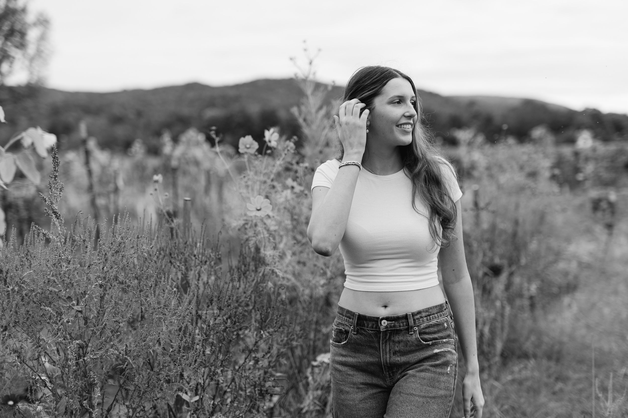 Black and white photo of someone in a white crop top and jeans walking through a wildflower field on a sunny day.