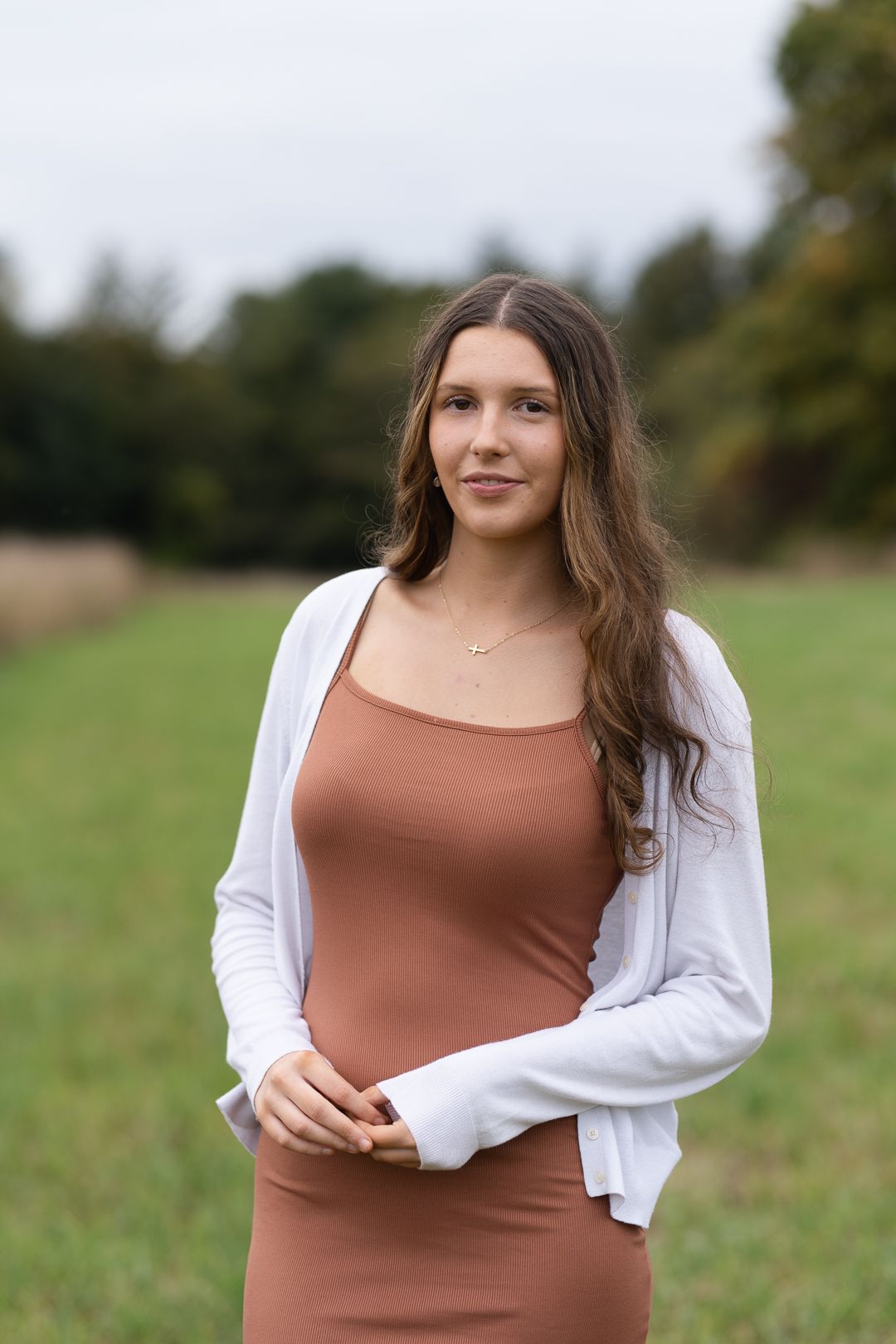 Person standing outdoors in a brown ribbed dress with white cardigan against a natural field background.