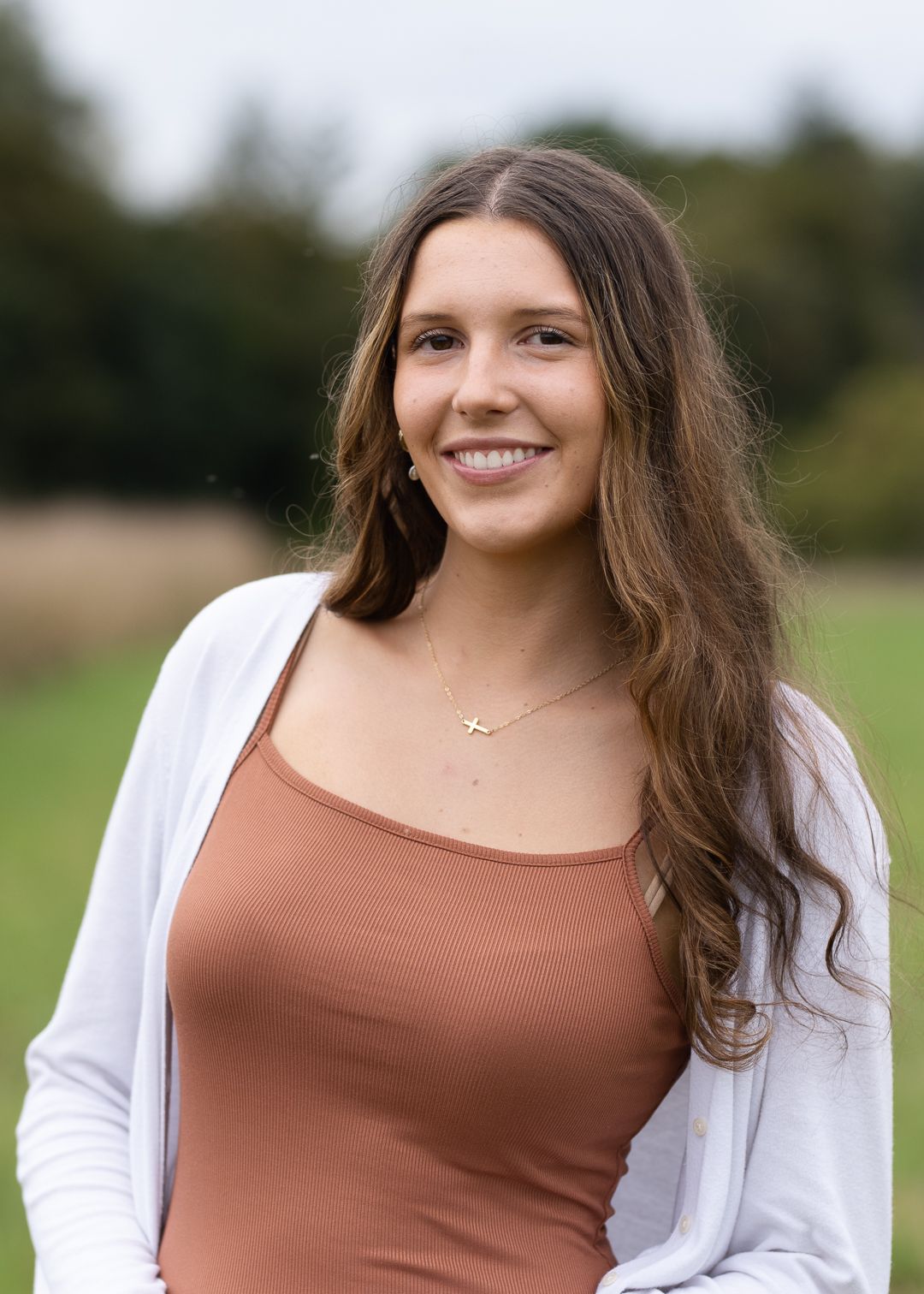 A person in a white cardigan and brown tank top stands outdoors against a natural blurred background.