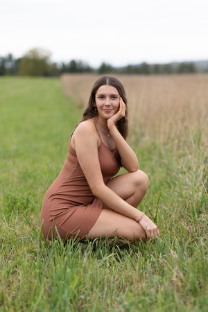 A person in a brown dress poses gracefully while sitting in a serene meadow field on a sunny day.