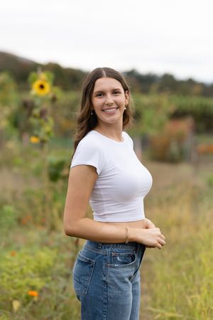 Person in white crop top and jeans poses in sunny field with sunflower in background.