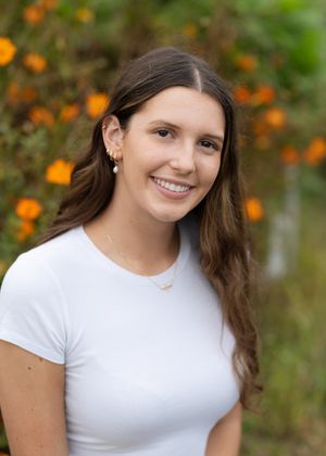 Young person in white t-shirt smiling outdoors with orange flowers blooming in the background.