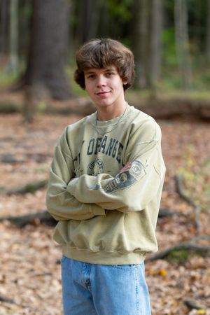 Young person wearing a tan collegiate sweatshirt and jeans poses with arms crossed in a forest setting.