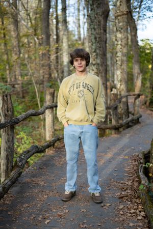 A person wearing a yellow sweatshirt and light jeans stands on a forest trail surrounded by tall trees.