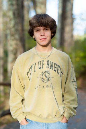 Young person in a light yellow 'City of Angels' sweatshirt poses outdoors in front of blurred trees.