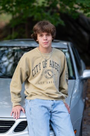 Person in a beige 'City of Angels' sweatshirt and light jeans leaning against car hood on a sunny day.