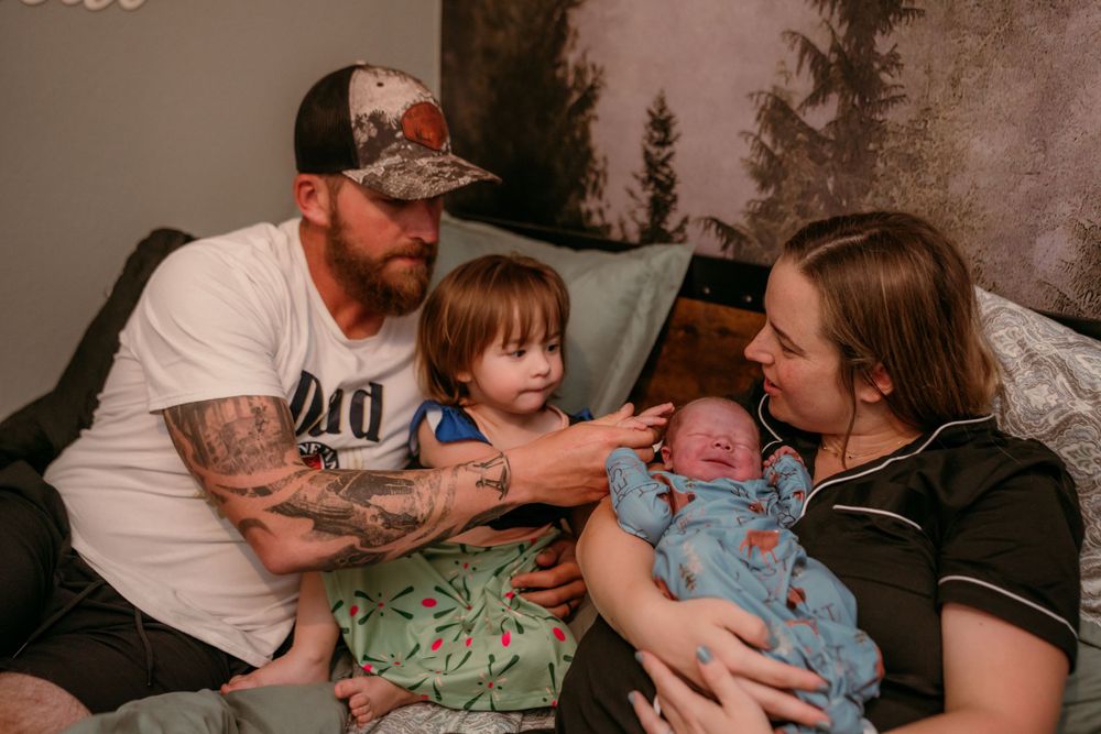 A family sits together on a couch, with a small child between two adults holding an infant sibling.