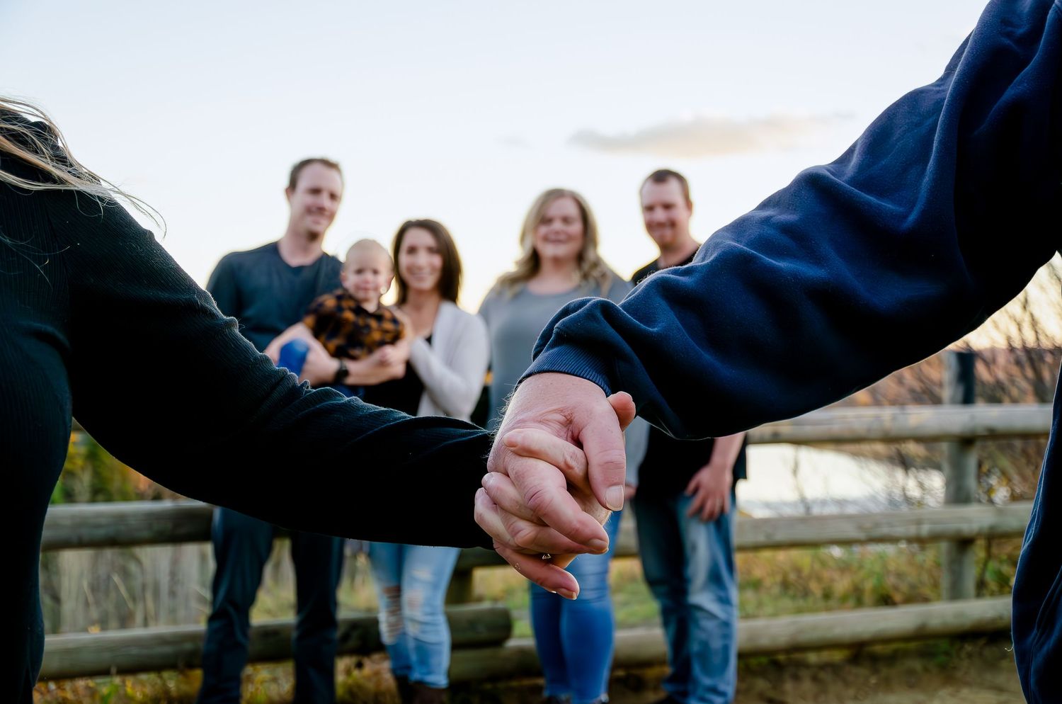 A group stands together outdoors with their hands joined in a circle at sunset near a wooden fence.