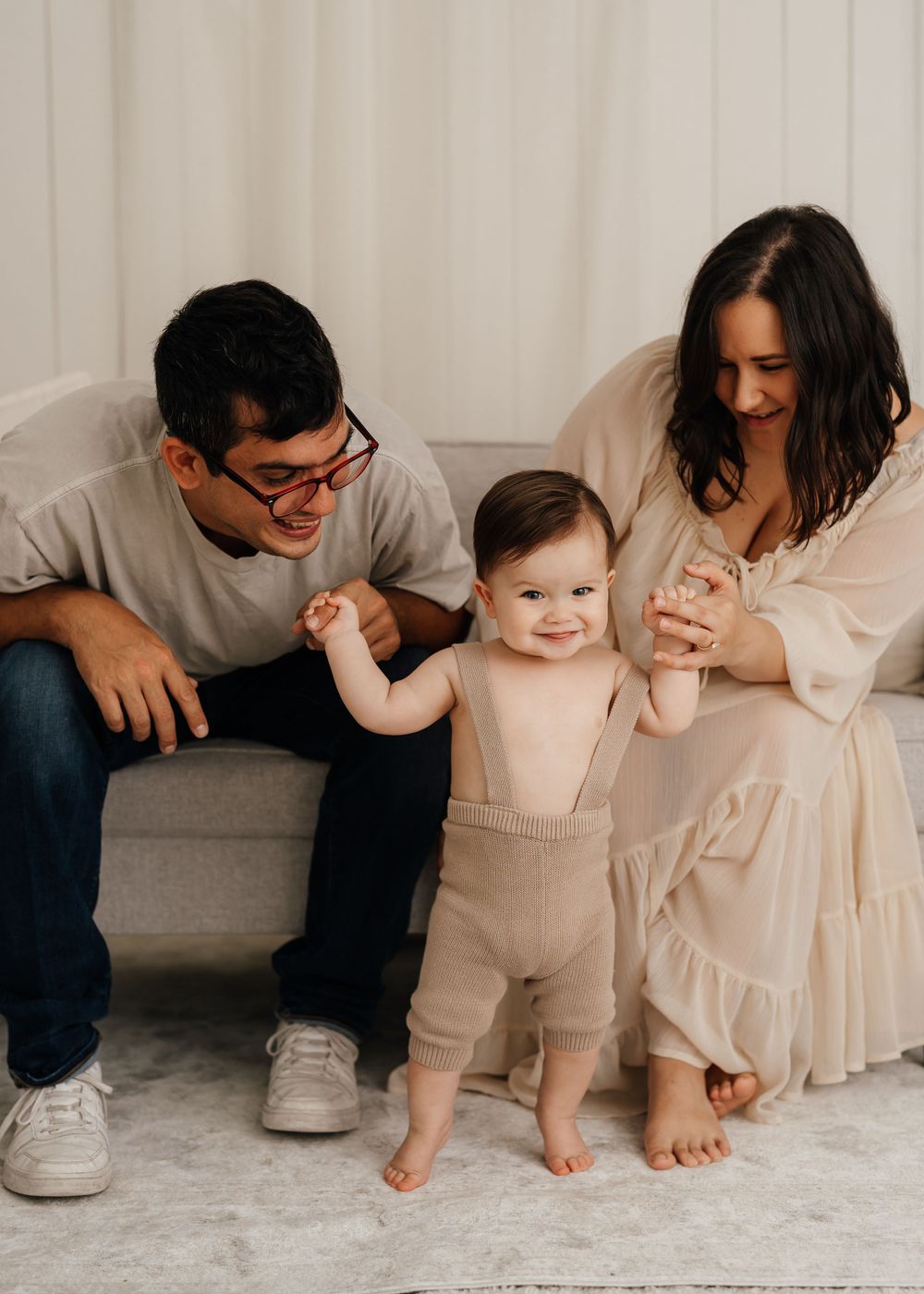 Loving family moment on couch as baby takes first steps in beige outfit while parents offer support.
