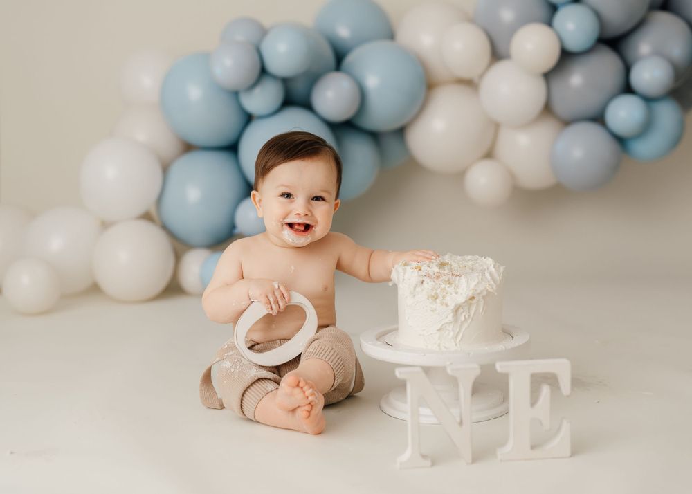 Joyful baby playing with smashed cake surrounded by blue and white balloon decorations.