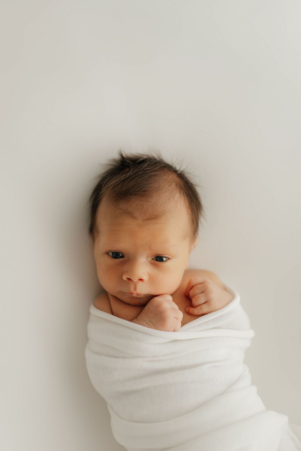 Newborn wrapped in white blanket against minimalist background.