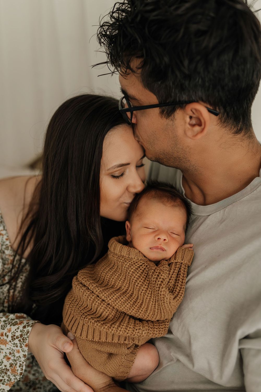 A tender family moment with a sleeping newborn in a brown knit sweater being cradled by parents.
