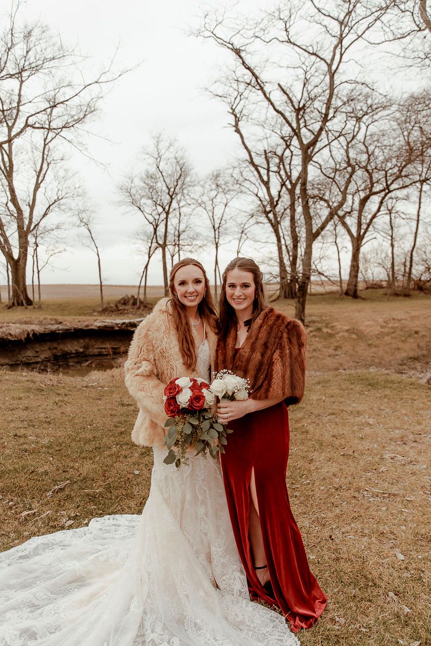 Two bridal attendants pose in a fall landscape wearing elegant gowns and fur wraps with a bouquet of red flowers.