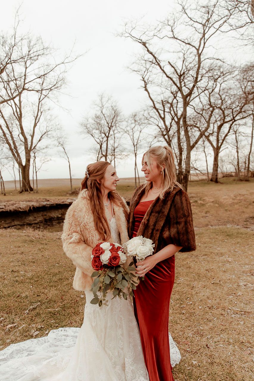 A winter wedding scene features two women in red velvet and white lace gowns standing among bare trees.