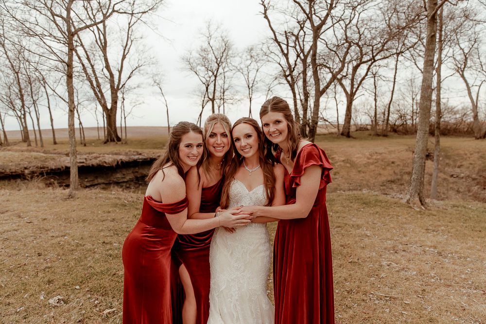 A group in burgundy dresses and white gown pose together outdoors on a fall day among bare trees.