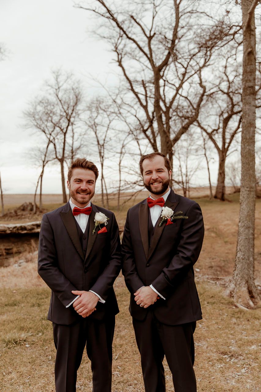 Two groomsmen in dark suits and bow ties pose together outdoors in front of bare trees on a cloudy day.