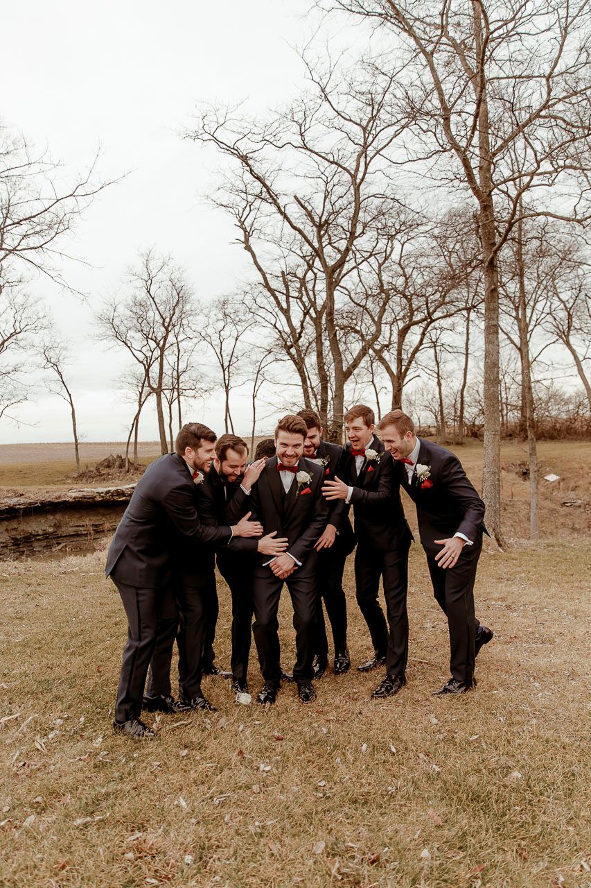 A group in black formal suits huddles together playfully outdoors against a backdrop of bare winter trees.