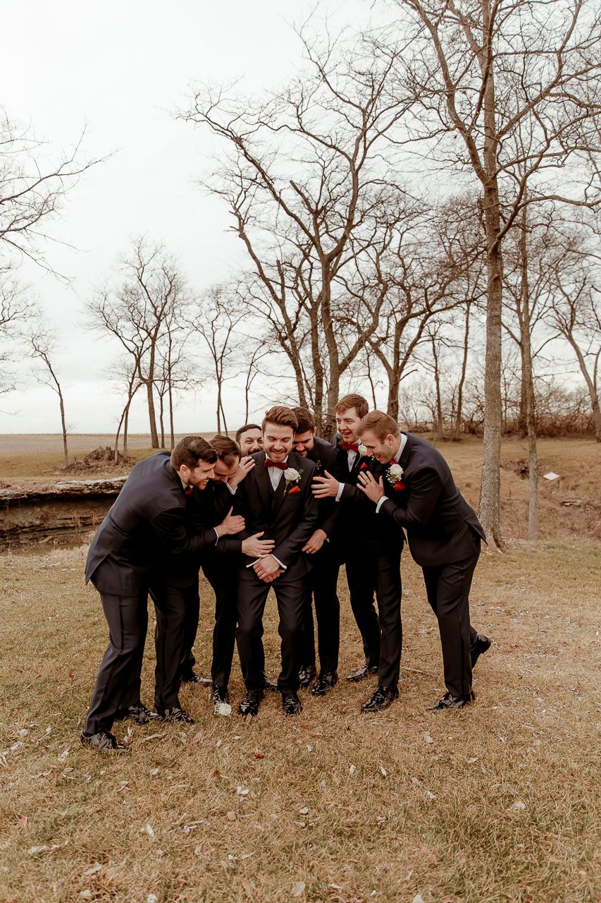 Groomsmen in black suits huddle together laughing in a playful pose against bare winter trees in autumn field.