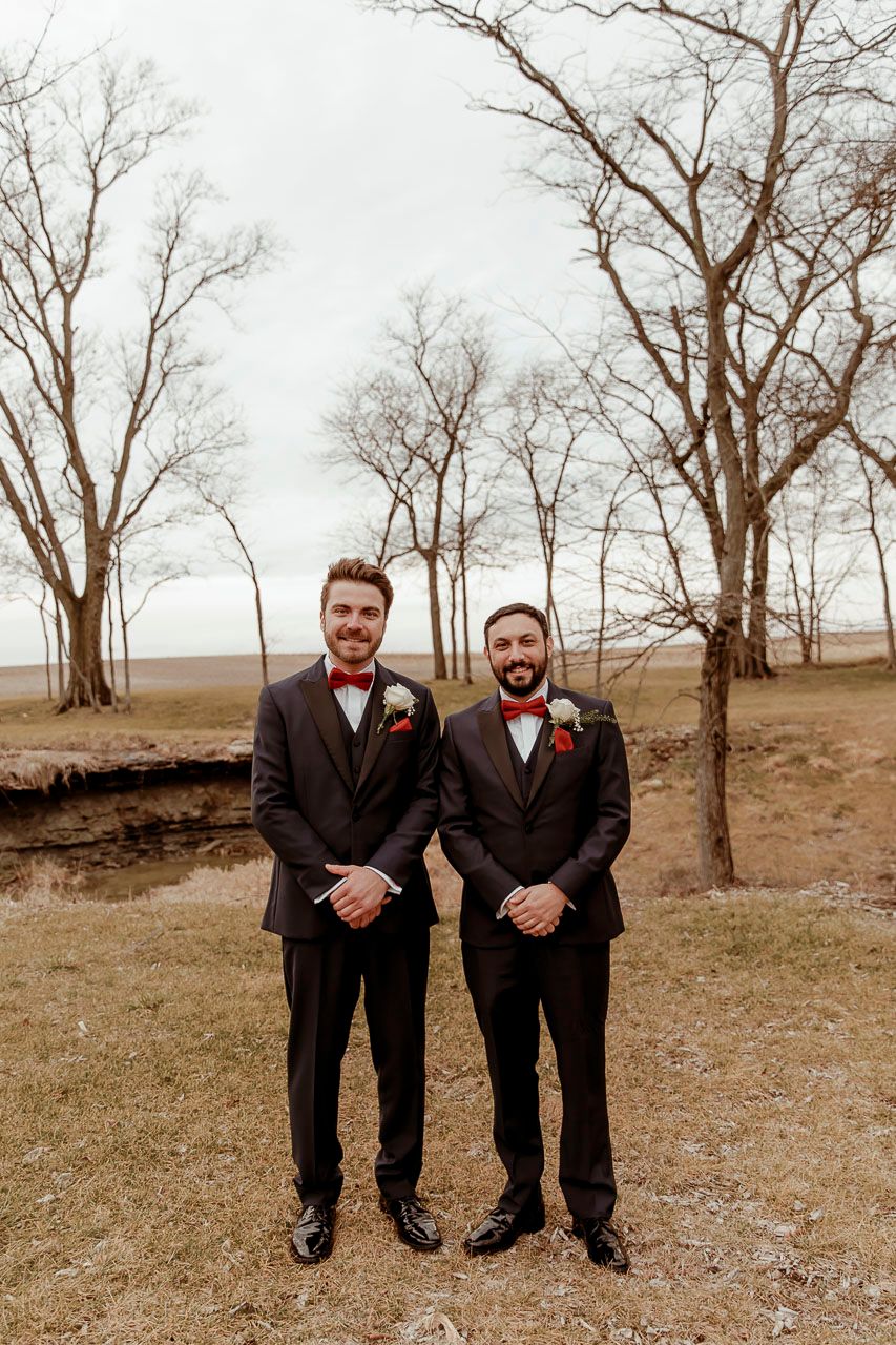 Wedding photograph featuring coordinated suits with red bow ties and boutonnieres against a backdrop of bare winter trees.