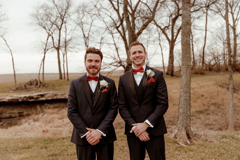 Two people in dark suits with red bow ties stand together in a wooded outdoor setting during autumn.