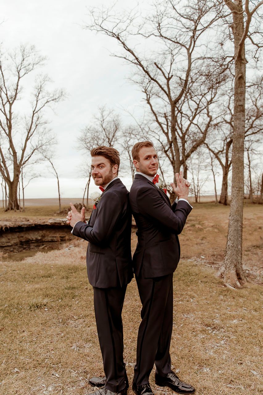 Two men in black suits pose back-to-back in a playful stance among bare trees in an outdoor setting.