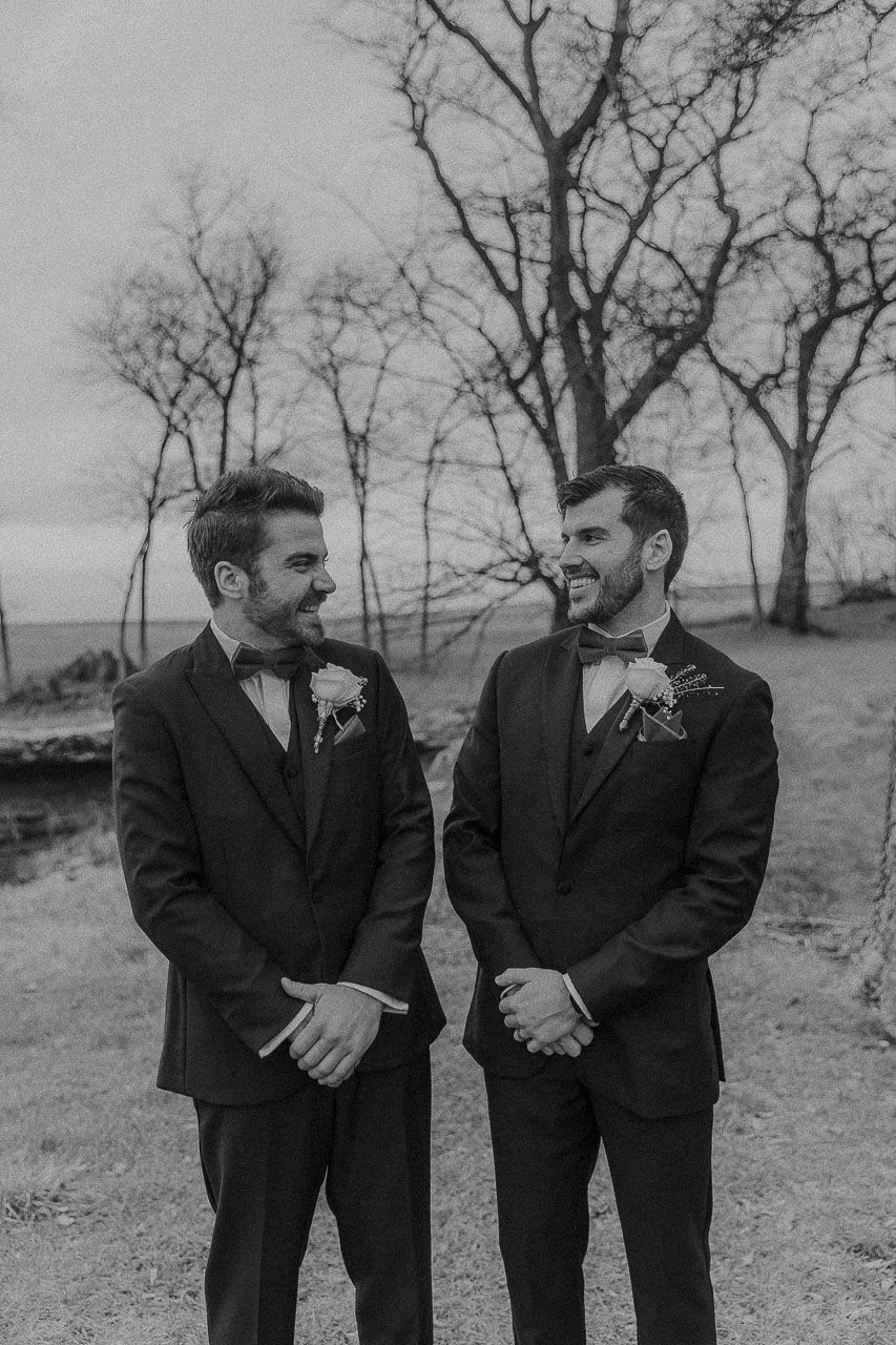 Black and white portrait of two people in formal suits with bow ties sharing a joyful moment outdoors.