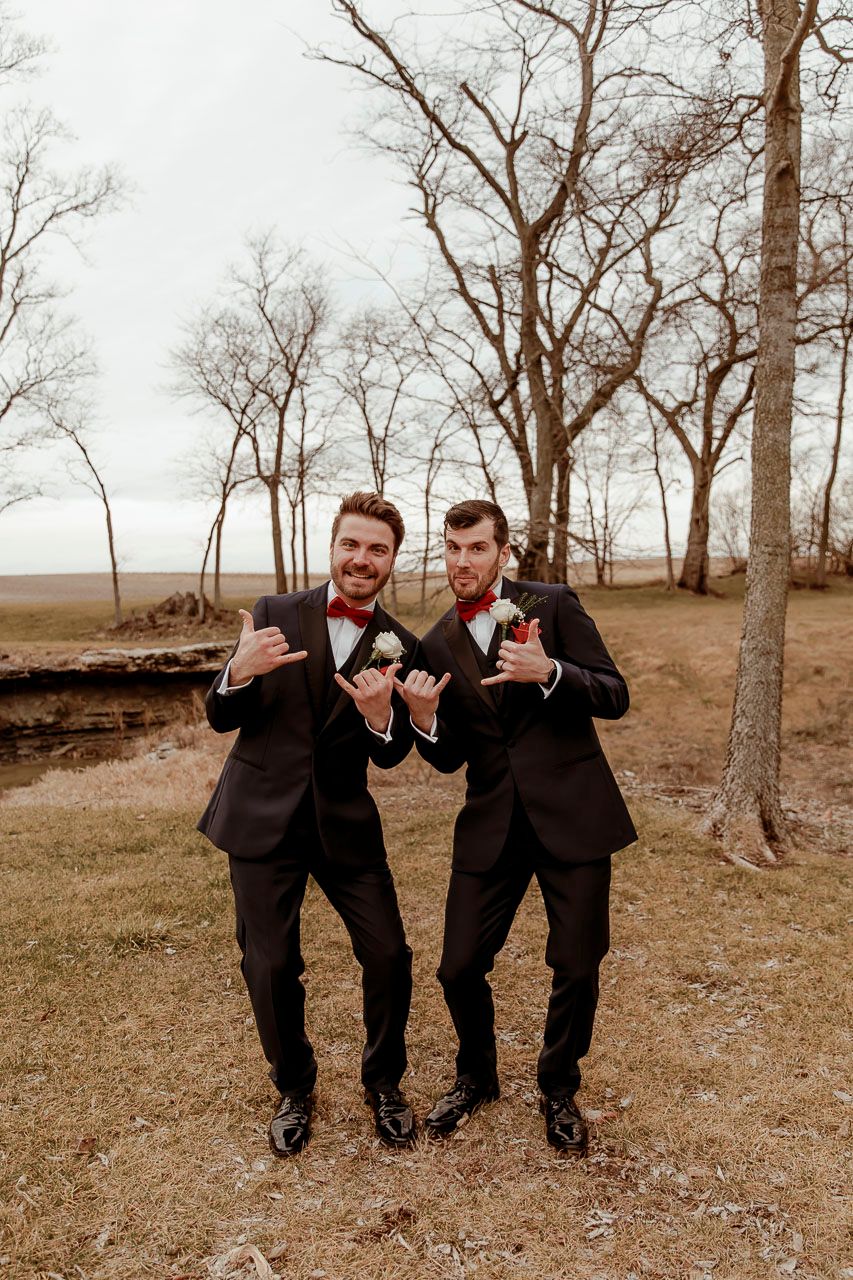 Two men in matching dark suits and bowties pose together outdoors among bare trees on a grassy landscape.