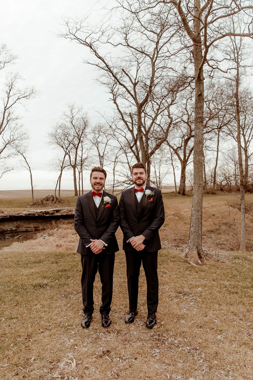 Two matching groomsmen in black tuxedos with red bowties stand among bare trees on autumn grounds.