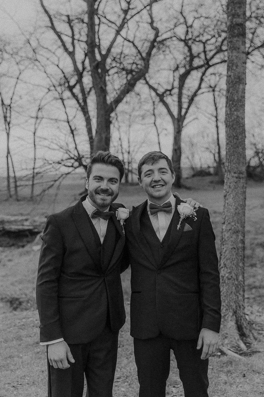 Two people wearing black tuxedos with bow ties pose together outdoors against a backdrop of bare winter trees.