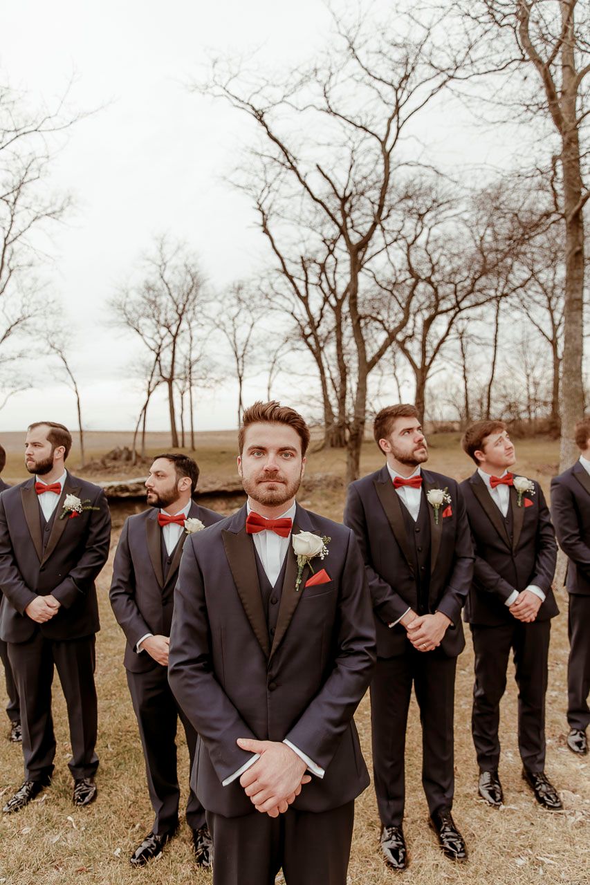 Group of groomsmen in black tuxedos and red bow ties stand in formation outdoors during a wedding photoshoot.