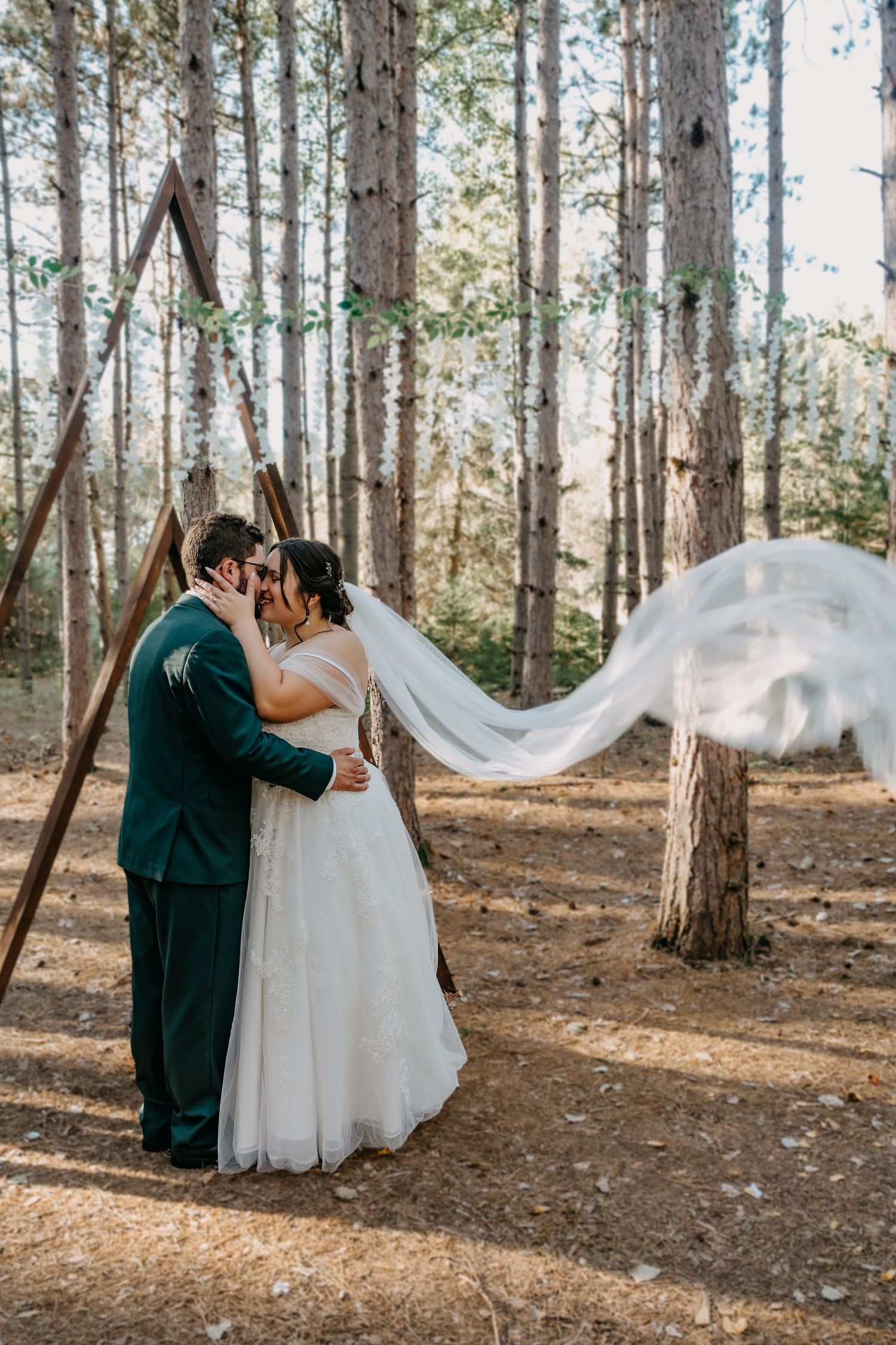 A romantic forest wedding ceremony with a wooden triangle arch and flowing white veil catching the breeze among tall pine trees.