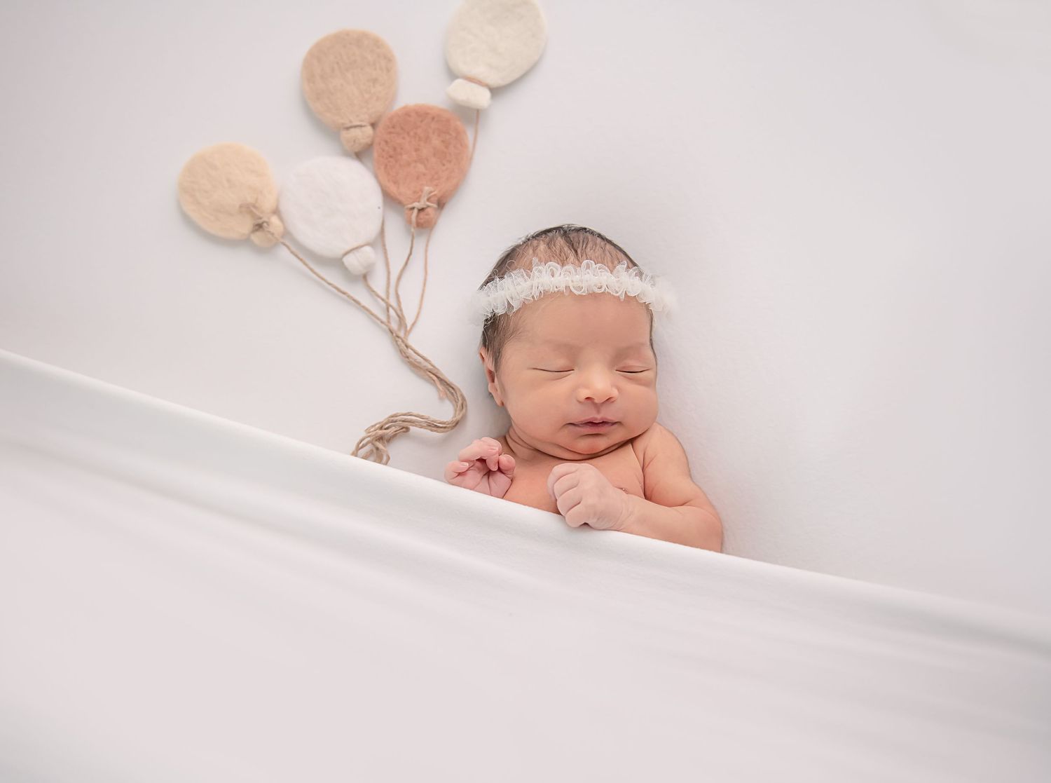 Sleeping newborn with white headband lying next to pale pink and beige balloon decorations.