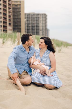 A couple sits together on a sandy beach with a baby, high-rise buildings visible in the background.