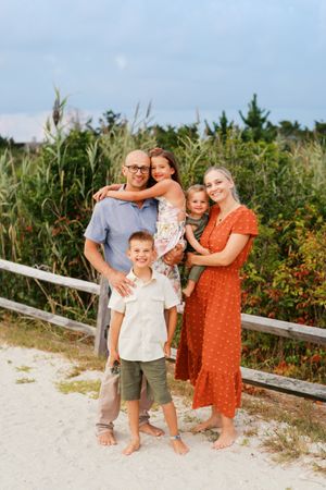 Family of five standing together on sandy beach path with tall grass and blue sky in background.