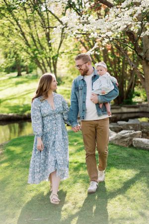 A family takes a springtime stroll together near a pond with blooming cherry blossoms overhead on a sunny day.