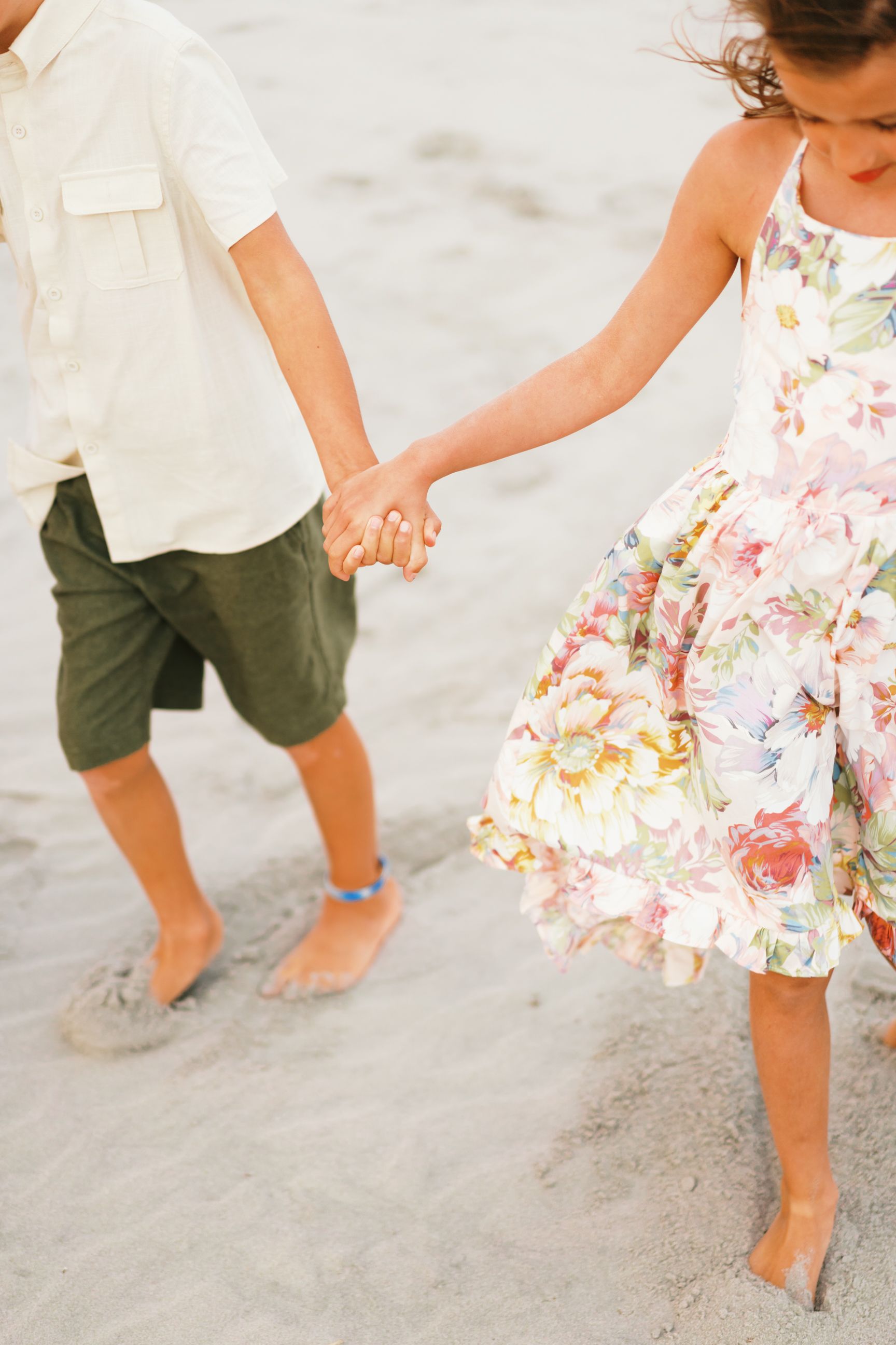 Couple holding hands while walking barefoot on beach, woman wearing floral summer dress and man in white shirt and shorts.