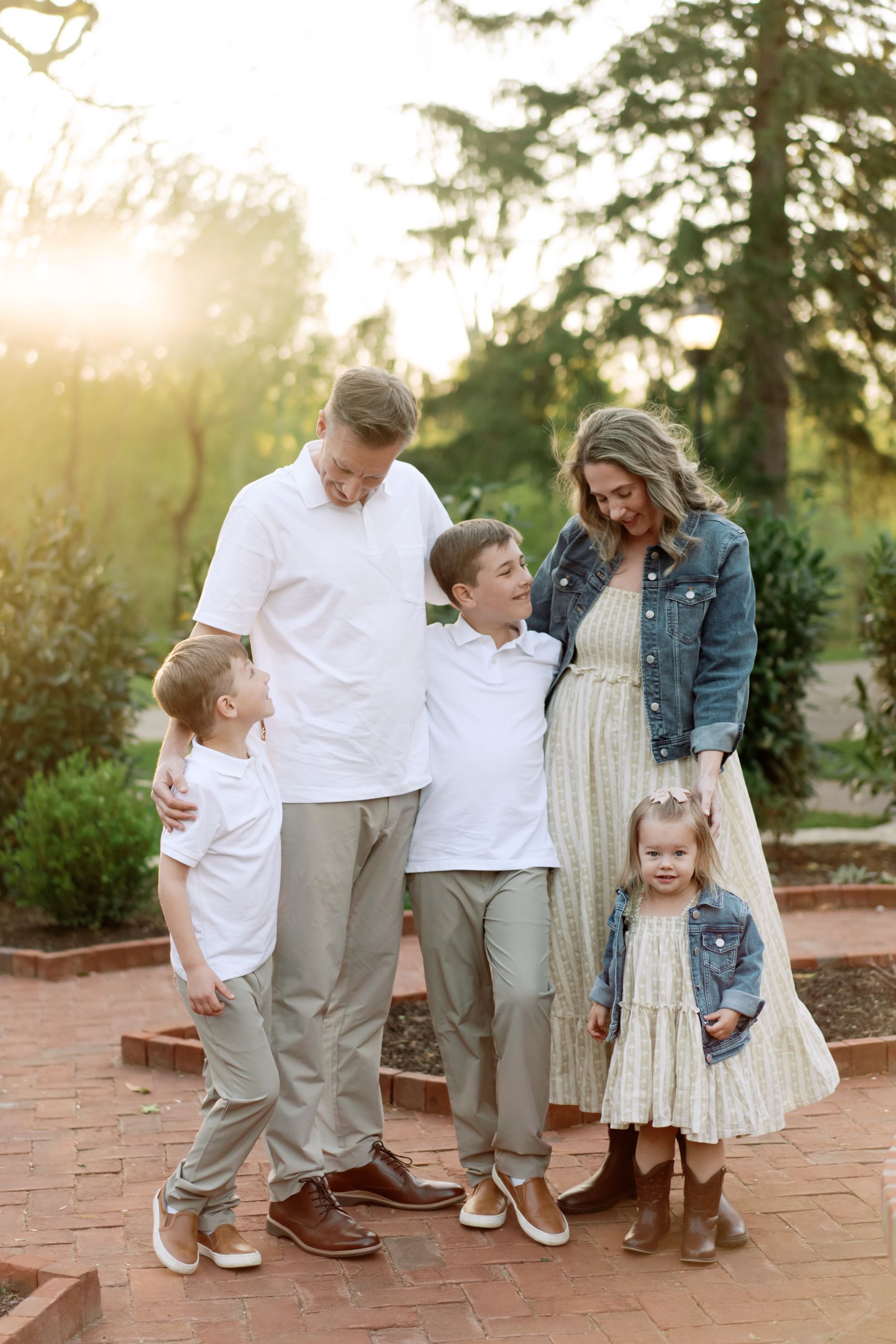 Family of five outdoors at sunset wearing coordinated white and beige outfits standing on brick path in garden setting.