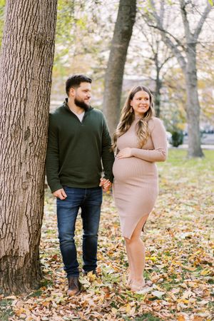 A couple holding hands while posing in an autumn park with fallen leaves on the ground.
