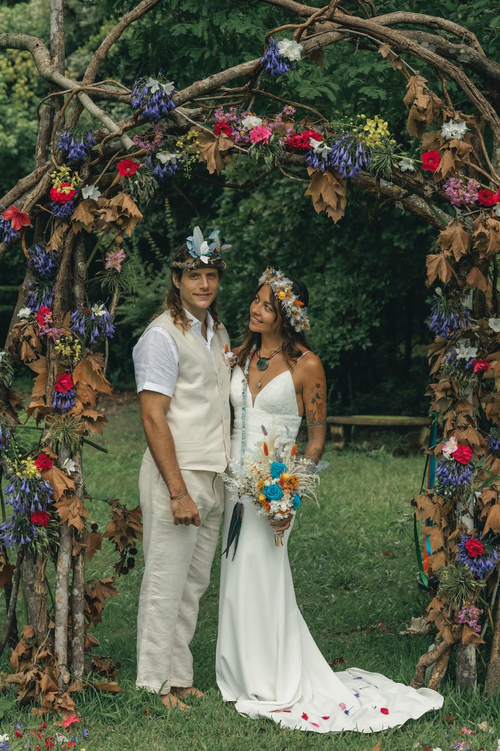 A wedding ceremony under a floral arch with red and blue flowers in a garden setting.