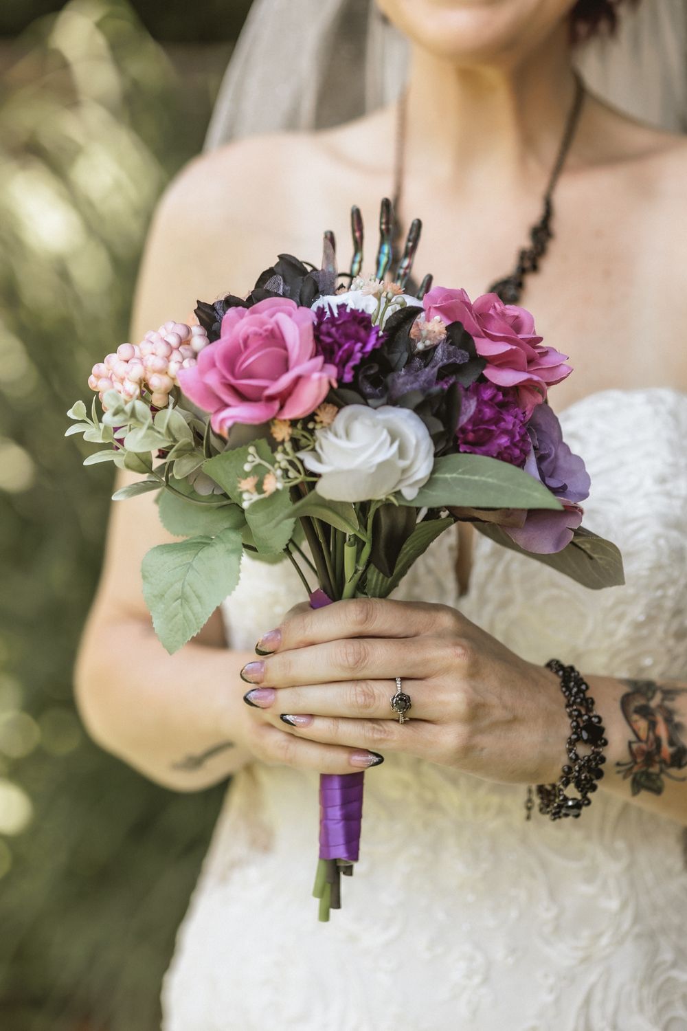 Wedding bouquet with purple and white roses wrapped in purple ribbon held by hands with dark nail polish.
