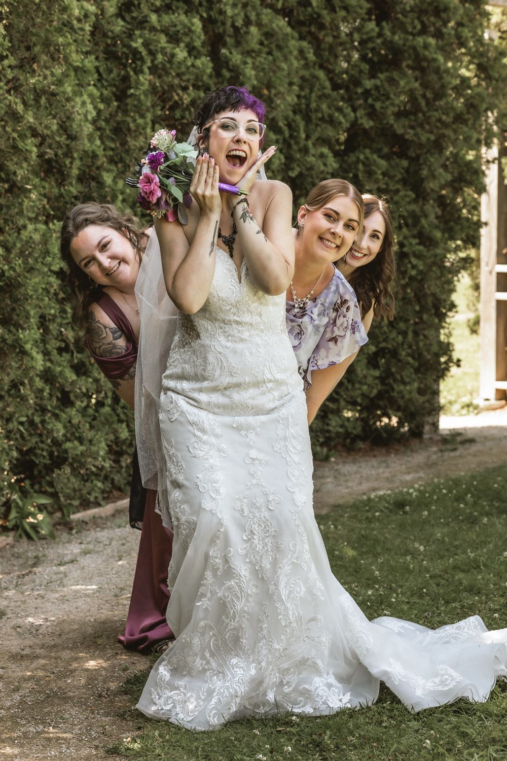 Wedding party poses playfully on grass with bride in white lace dress at center.