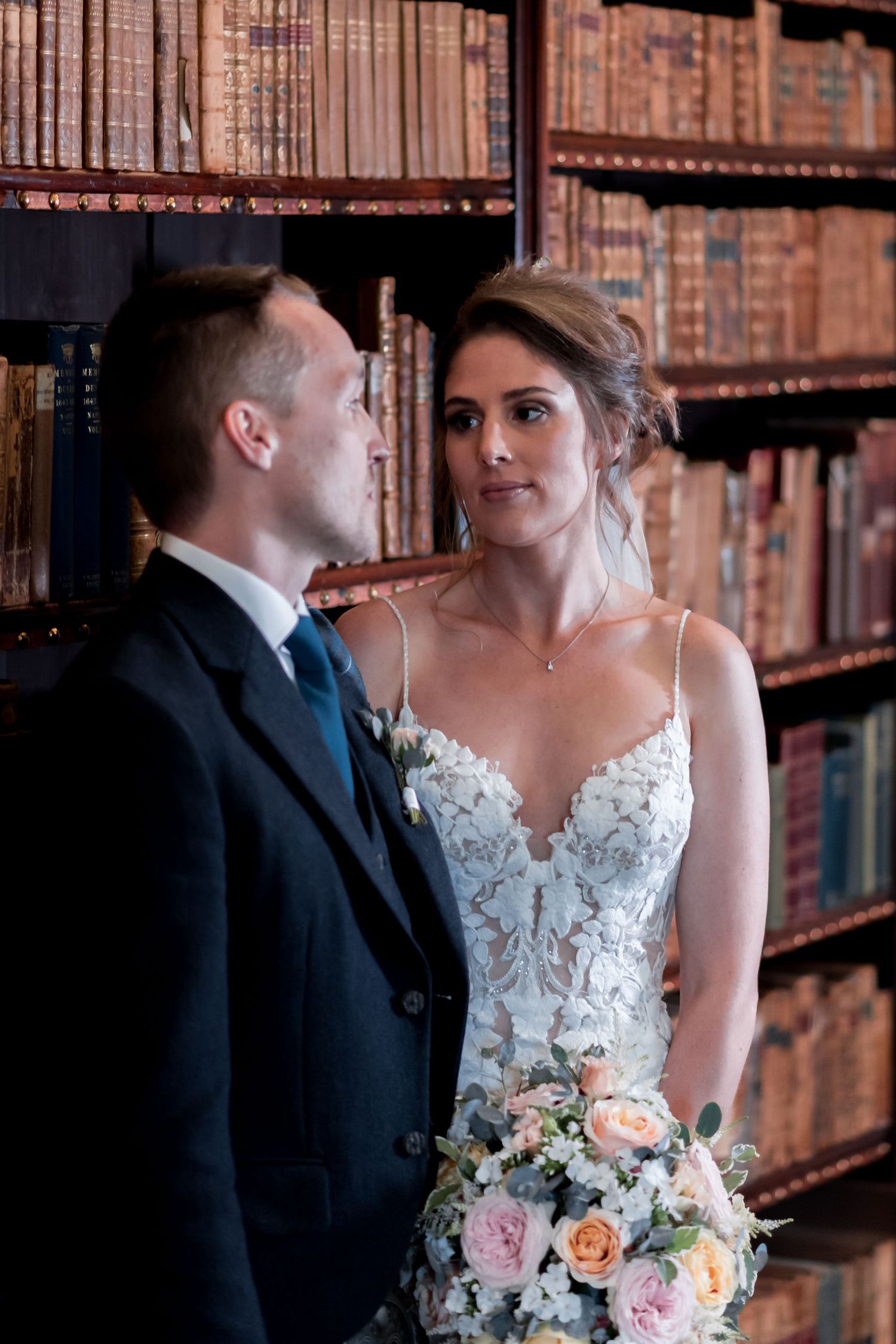 A bride and groom share an intimate moment in a library with vintage books and a pastel bouquet in the frame.