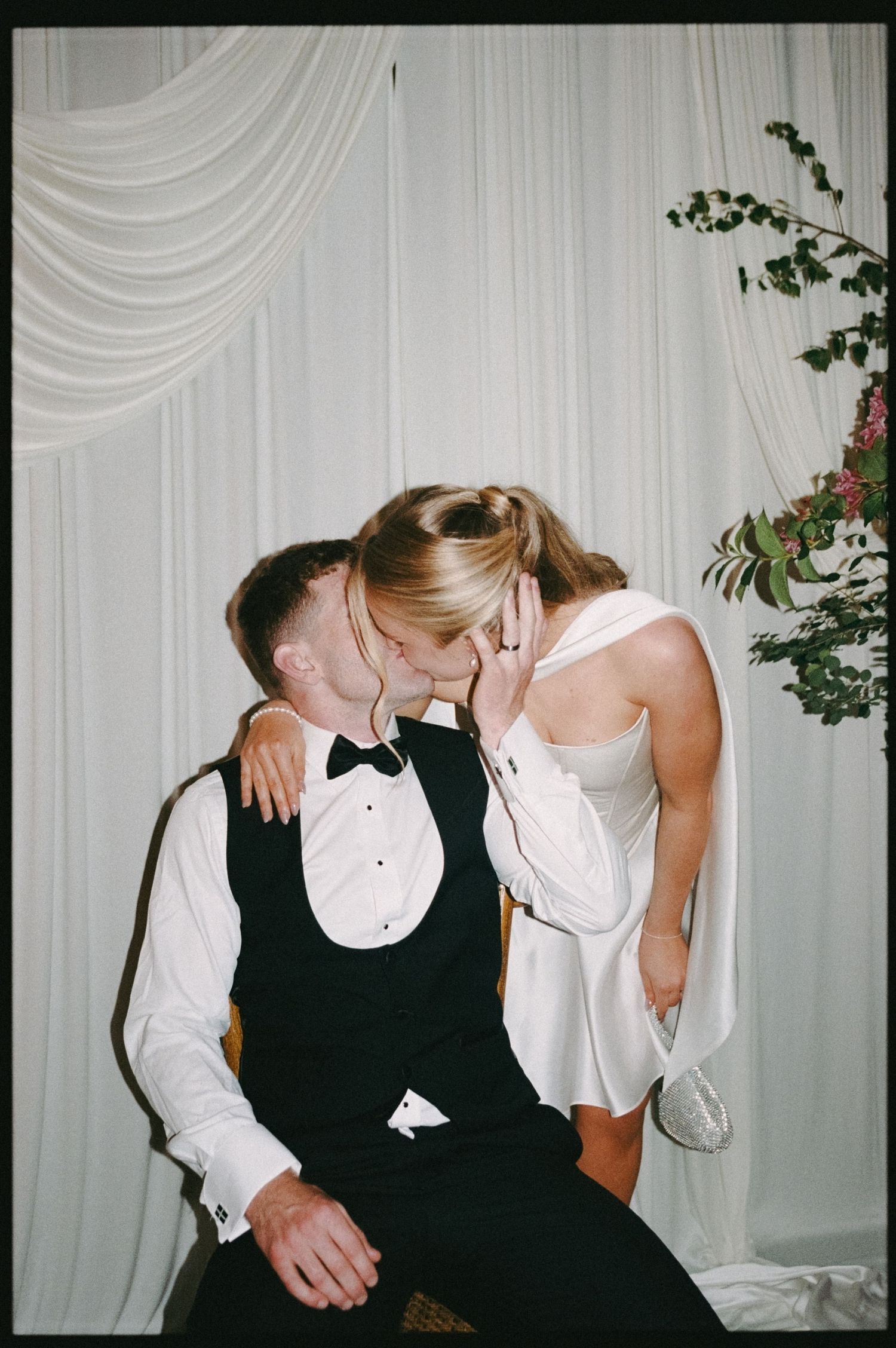 A bride and groom share a romantic kiss at their wedding reception, framed by elegant white draping and floral arrangements.