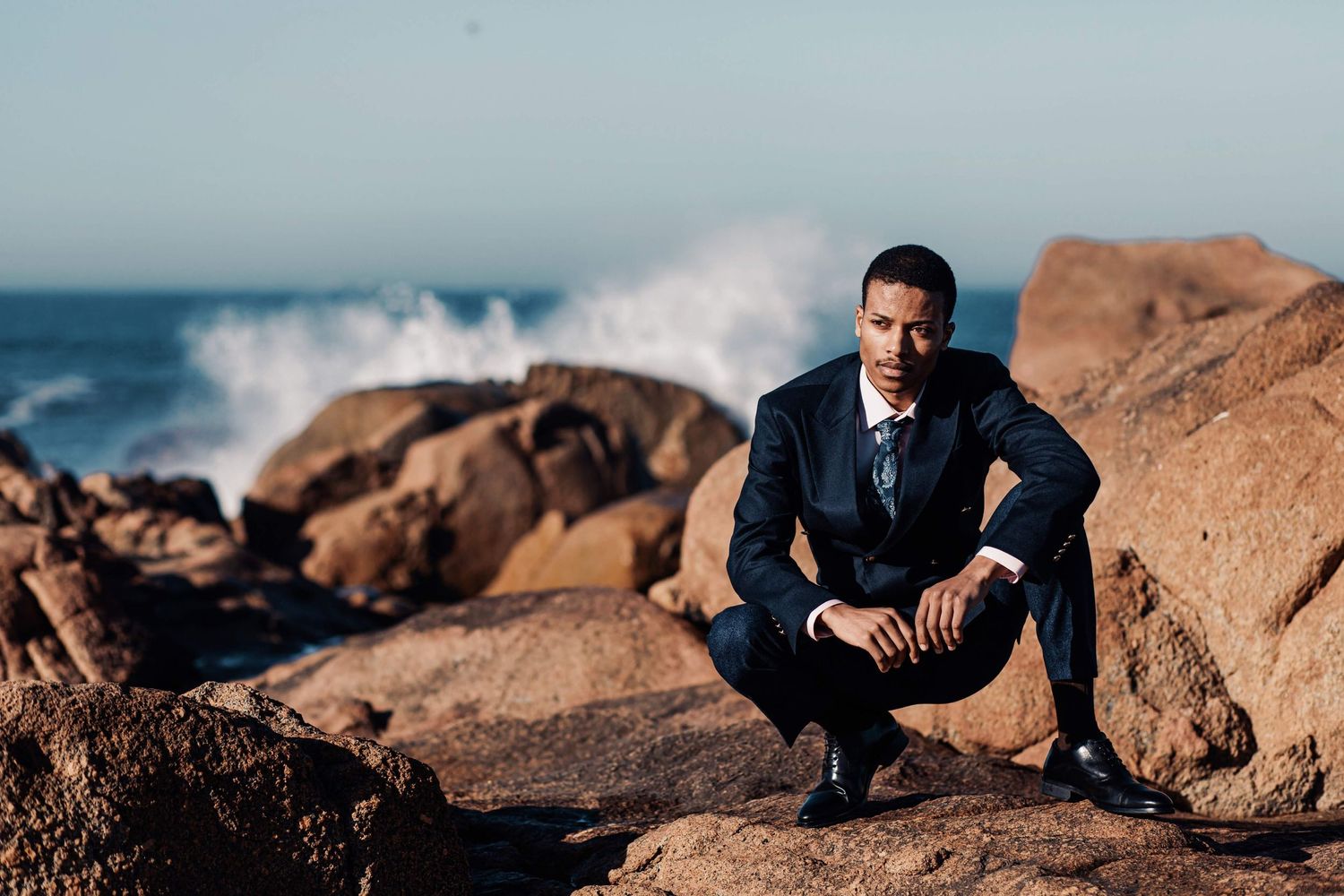 Someone in a dark suit crouches contemplatively on coastal rocks as ocean waves crash behind them at sunset.